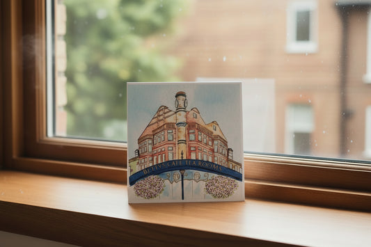 A square printed card or art print featuring a painted illustration of the Betty's Coffee Tea Rooms building, with a blue banner across the front and purple flower bouquets at the bottom, photographed standing on a white surface against a neutral beige background.