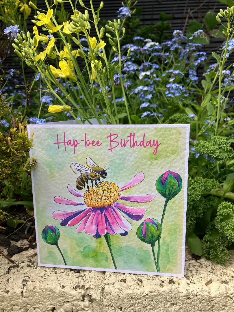 A square birthday card featuring a watercolor illustration of a bee on a pink flower, with the text 'Hap-bee Birthday' in pink script. The card is displayed outdoors on a stone surface surrounded by green plants and yellow flowers.