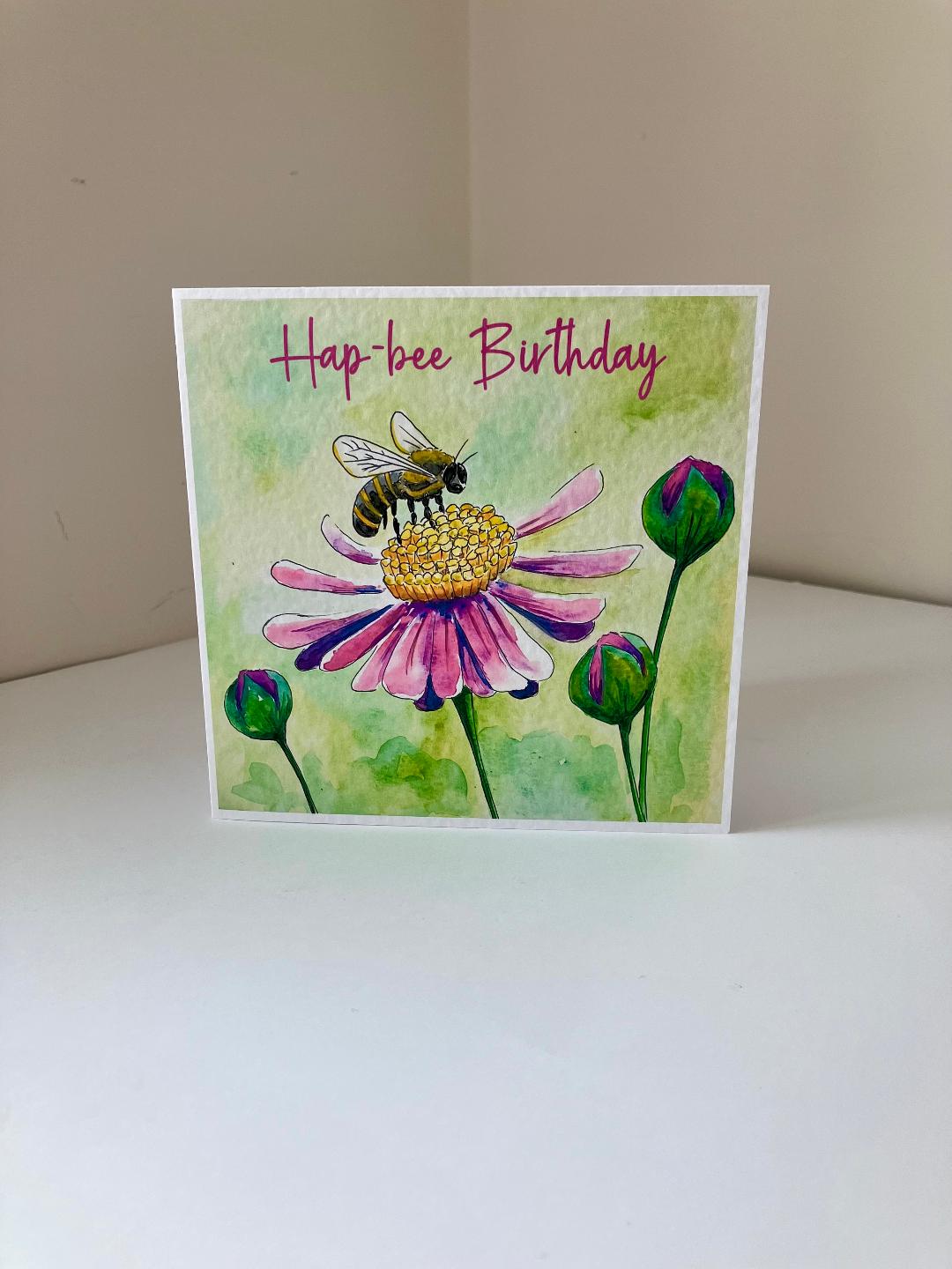 A square greeting card with a watercolor-style illustration of a bee on a pink flower, with the text 'Hap-bee Birthday' at the top, photographed standing on a white surface against a beige wall.