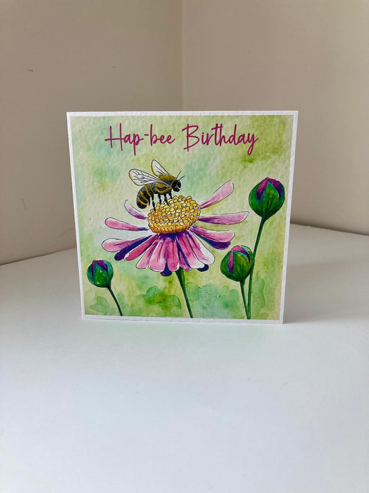 A square greeting card with a watercolor-style illustration of a bee on a pink flower, with the text 'Hap-bee Birthday' at the top, photographed standing on a white surface against a beige wall.