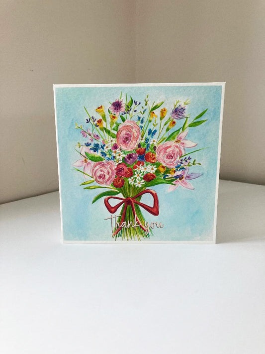 A square greeting card featuring a colorful watercolor bouquet of flowers tied with a red ribbon and the words 'Thank you' in cursive, displayed on a white surface against a beige background.